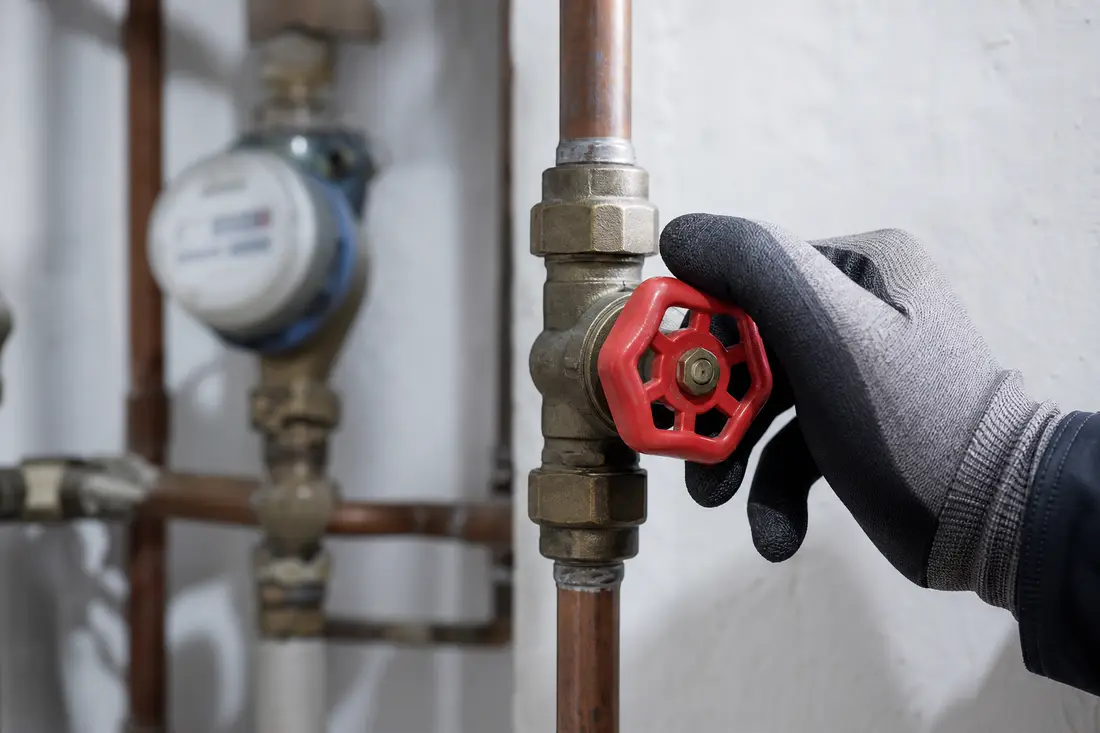 Hand in Arbeitshandschuh dreht den Hauptabsperrhahn einer Wasserleitung im Keller eines Wohnhauses zu